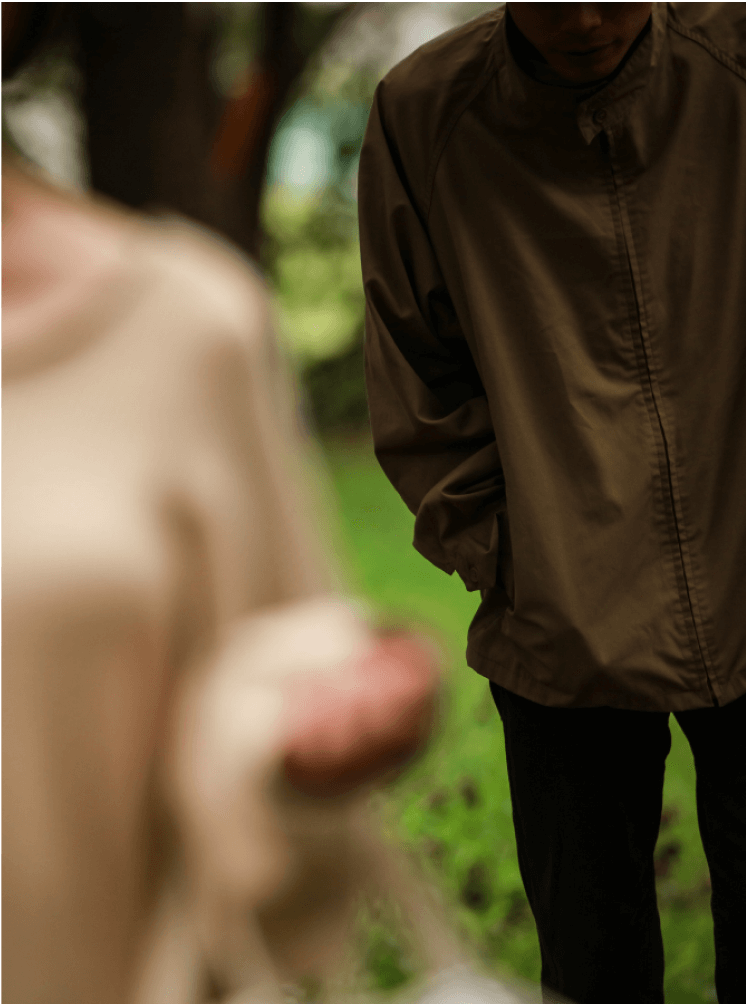 A person in a brown jacket standing outdoors with greenery in the background.