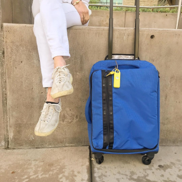 Person sitting beside a blue rolling suitcase with a yellow luggage tag.