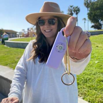Woman holding a keychain with a purple device in a sunny park.