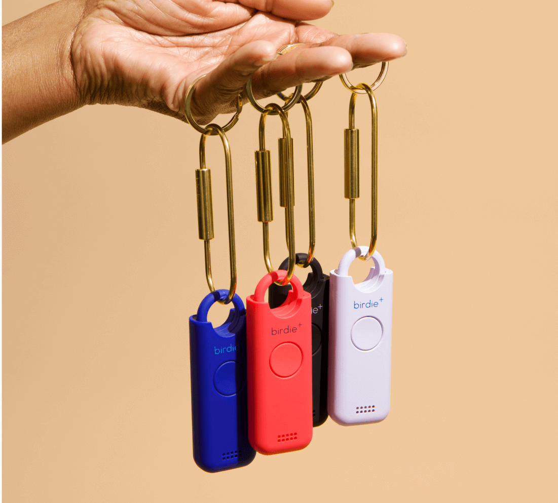 Hand holding three colorful personal safety alarms on keychains against a beige background.