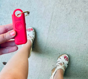 Hand holding a red personal safety alarm with white sandals in the background.