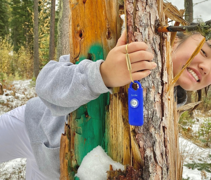 Smiling person peeking around a tree, holding a blue personal safety alarm.